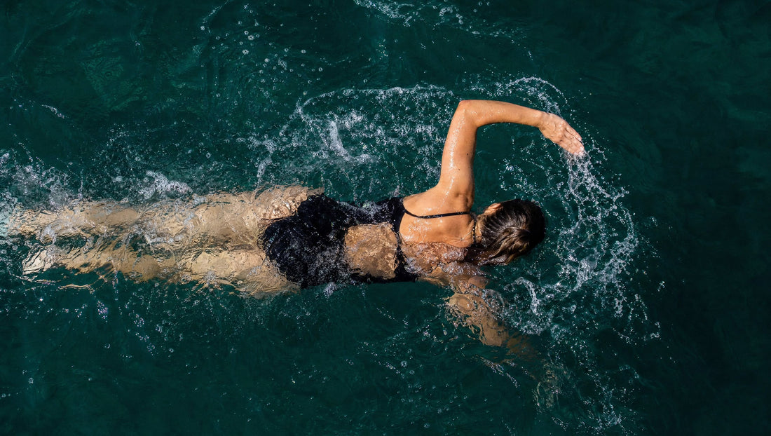 Frau schwimmt von oben gesehen im türkisgrünen Meer, ein Arm durchbricht die Wasseroberfläche