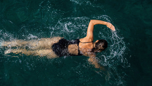 Frau schwimmt von oben gesehen im türkisgrünen Meer, ein Arm durchbricht die Wasseroberfläche