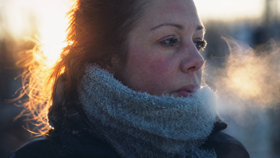 Frau mit Raureif auf Schal und Wimpern atmet in der Frostluft aus, Atemwolke im Gegenlicht